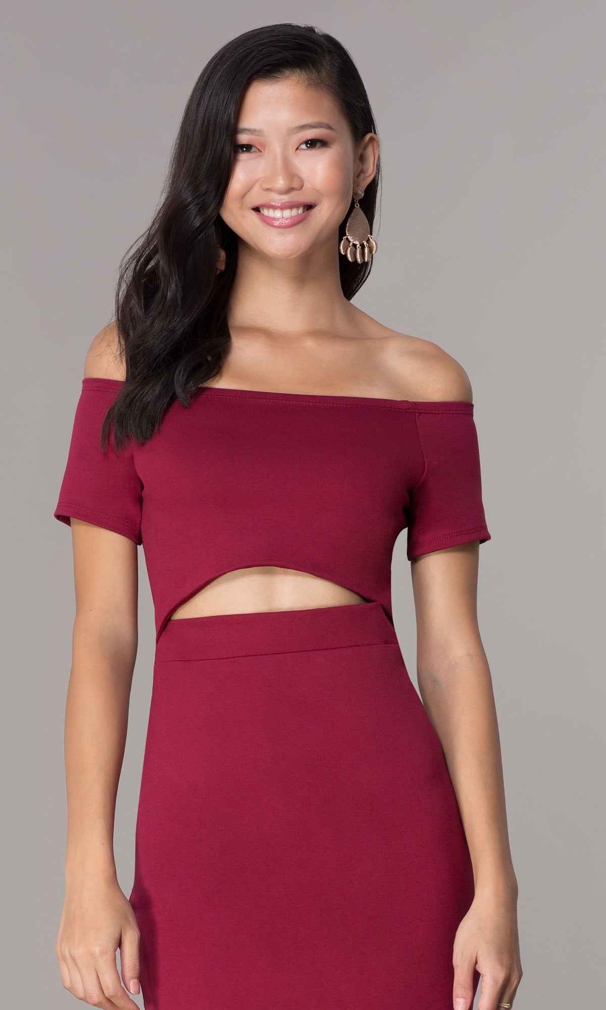 A woman smiles and poses in a burgundy Off-the-Shoulder Short Cut-Out Party Dress 3076 with short sleeves and a midriff cutout, set against a plain gray background.